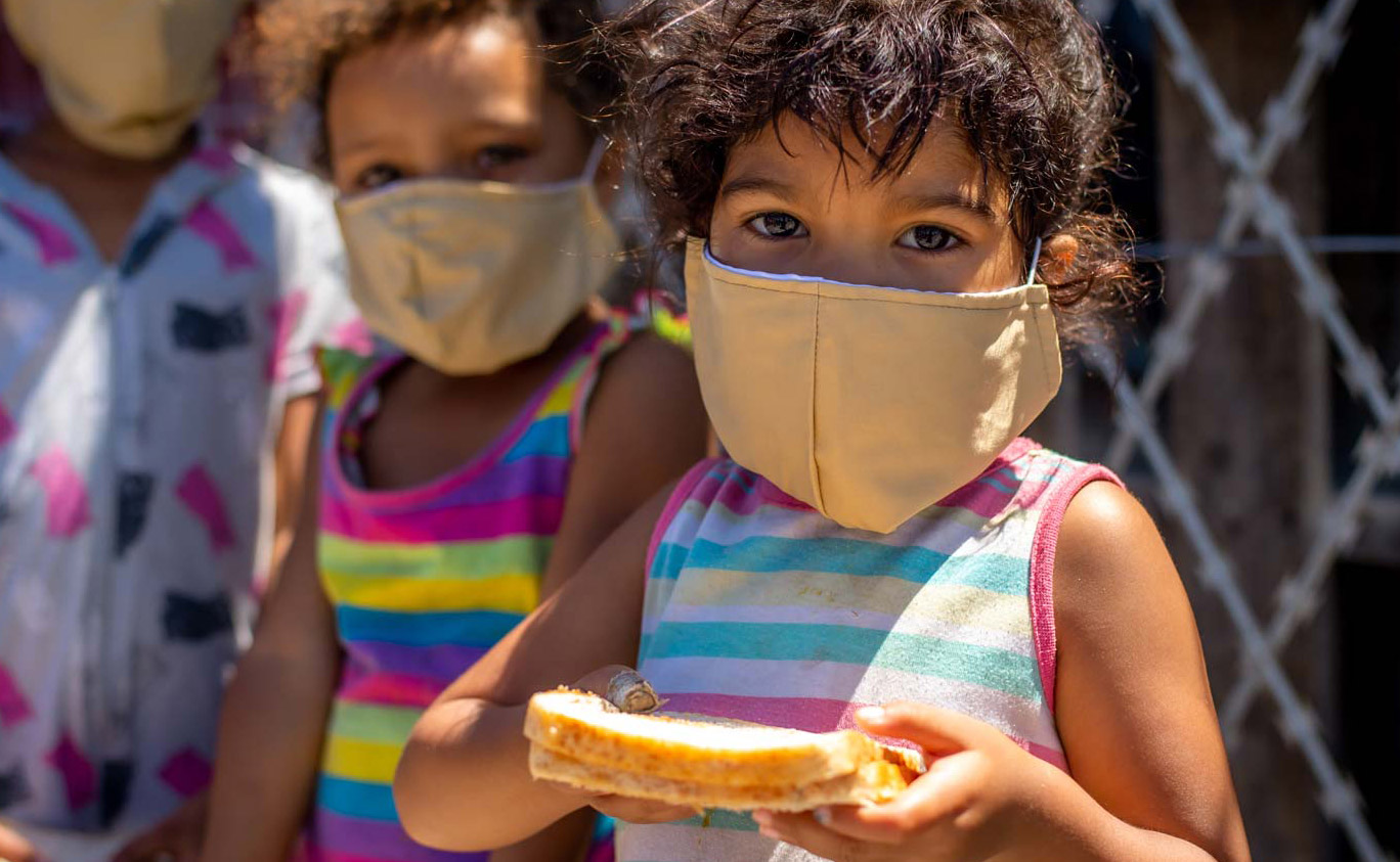 Small child at a soup kitchen wearing a facemask and holding a sandwich