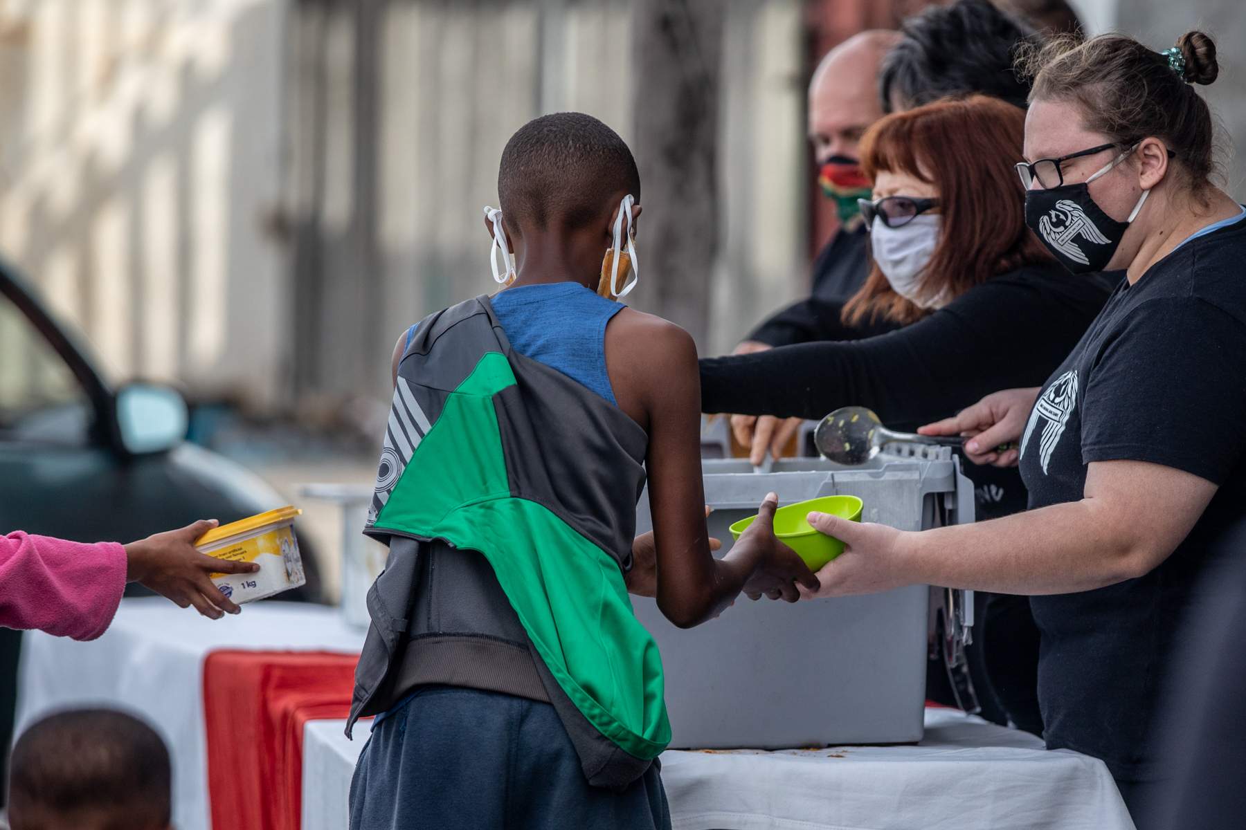 Boy with back to camera receives fresh soup from masked volunteers