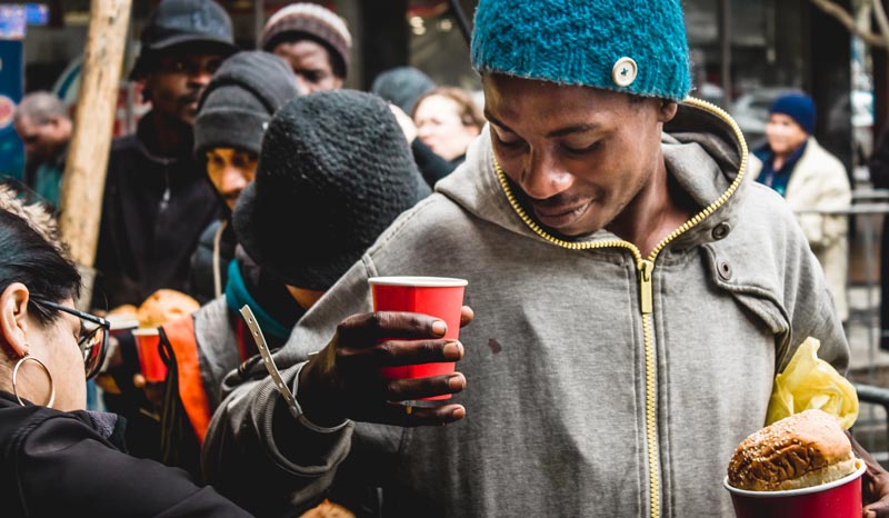 Guest at a soup kitchen smiles as he receives a bowl of soup, a bread roll and a cup of juice