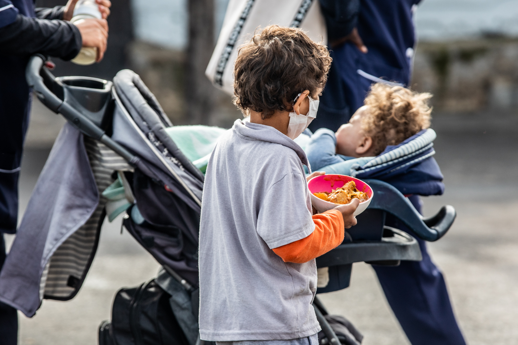 A boy holds a bowl of food as he walks next to a baby in a stroller